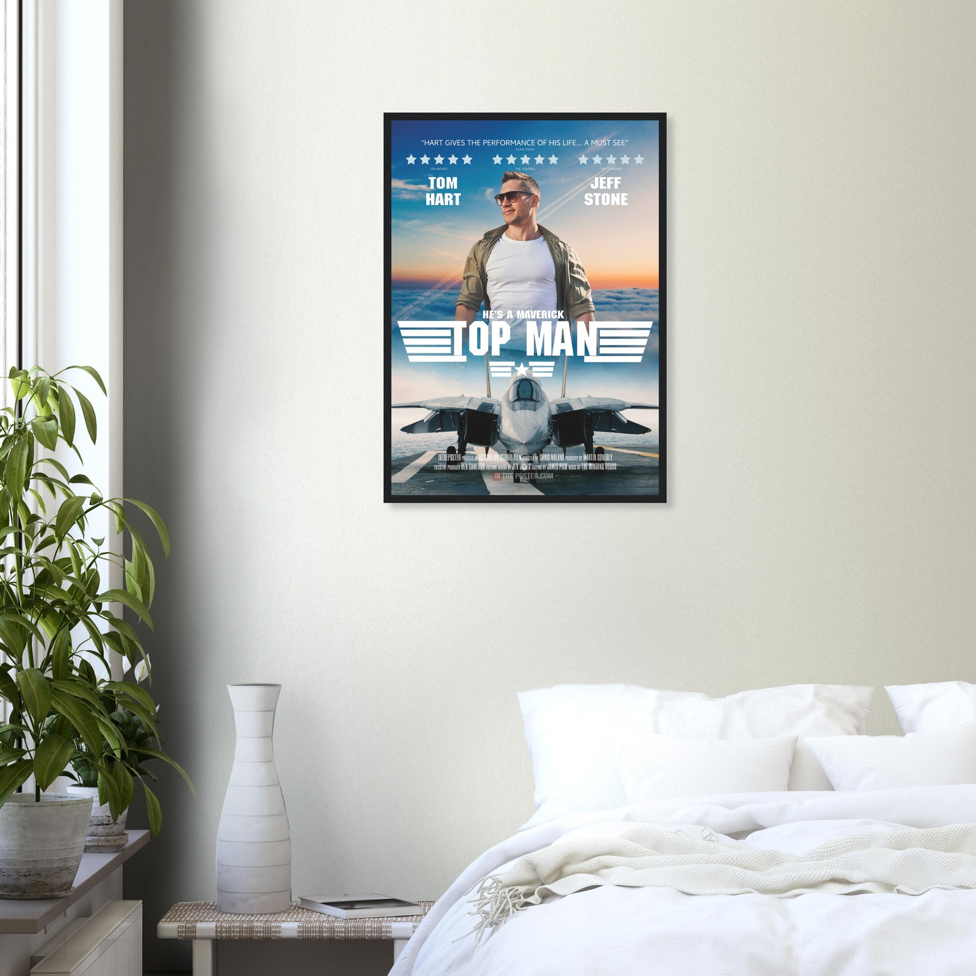 A fighter jet movie poster in regular size and a black frame, on a grey wall above a bed and green house plant