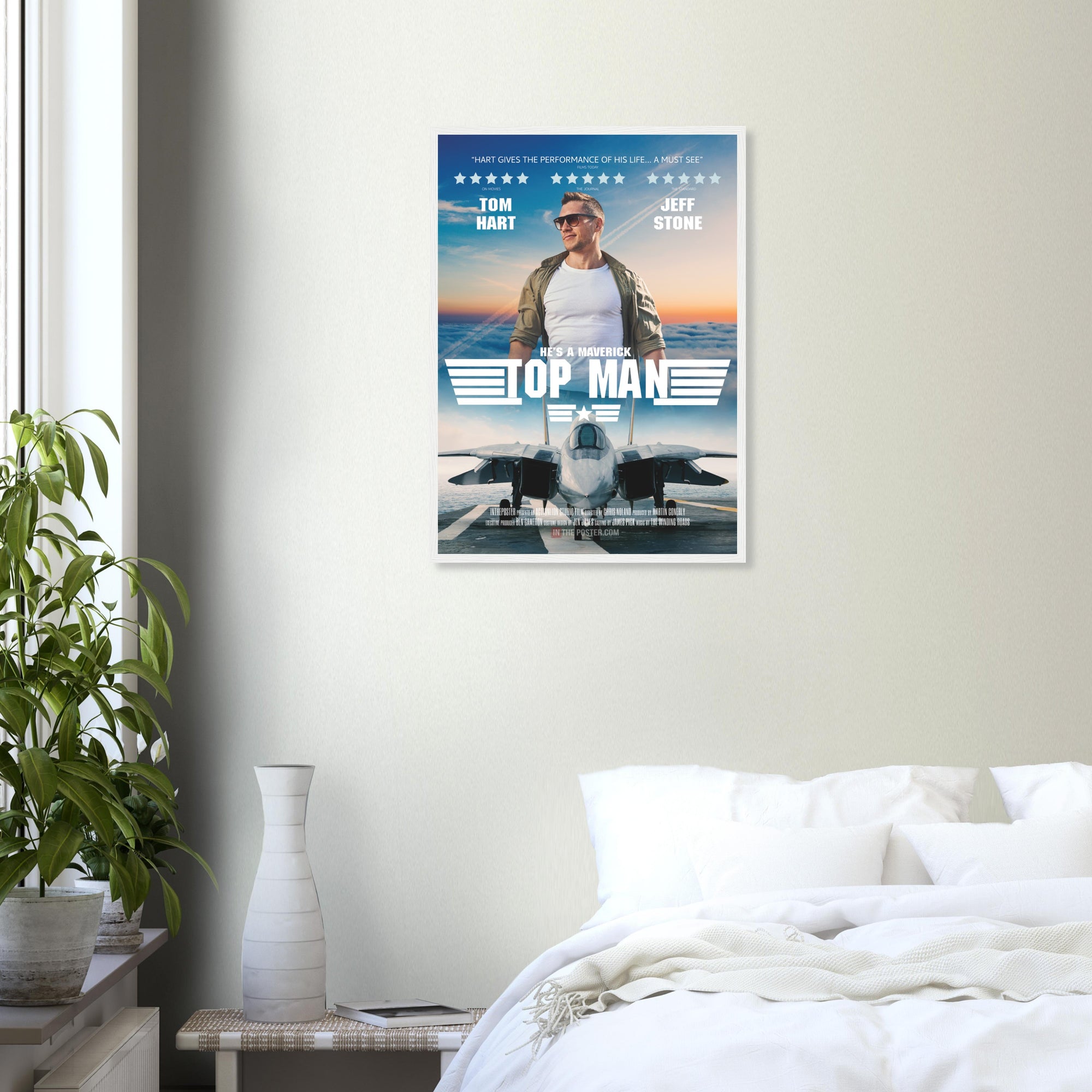 A fighter jet movie poster in regular size and a white frame, on a grey wall above a bed and green house plant