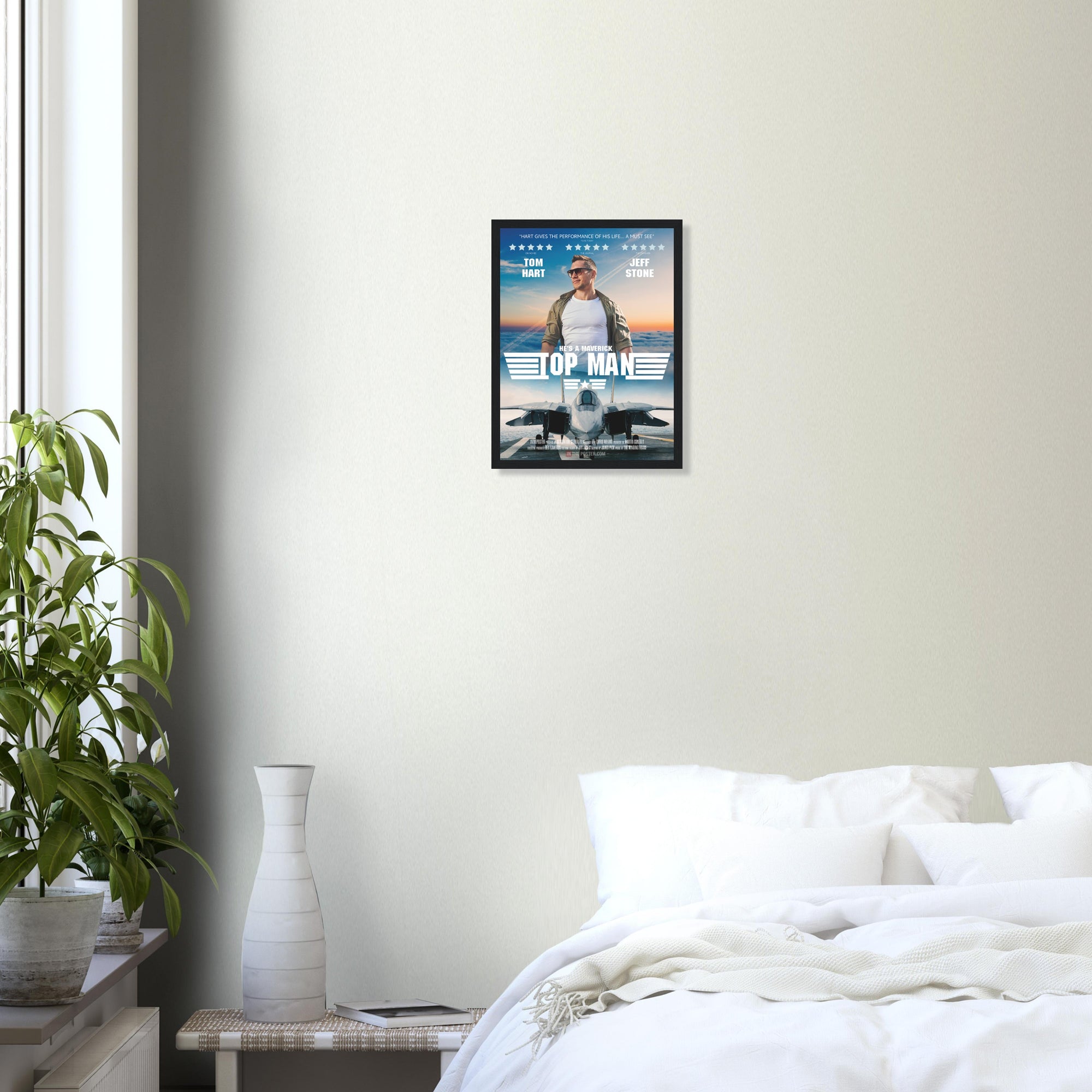 A fighter jet movie poster in small size and a black frame, on a grey wall above a bed and green house plant