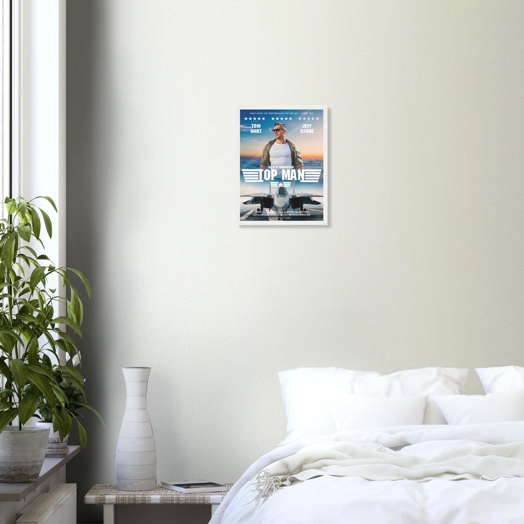 A fighter jet movie poster in small size and a white frame, on a grey wall above a bed and green house plant